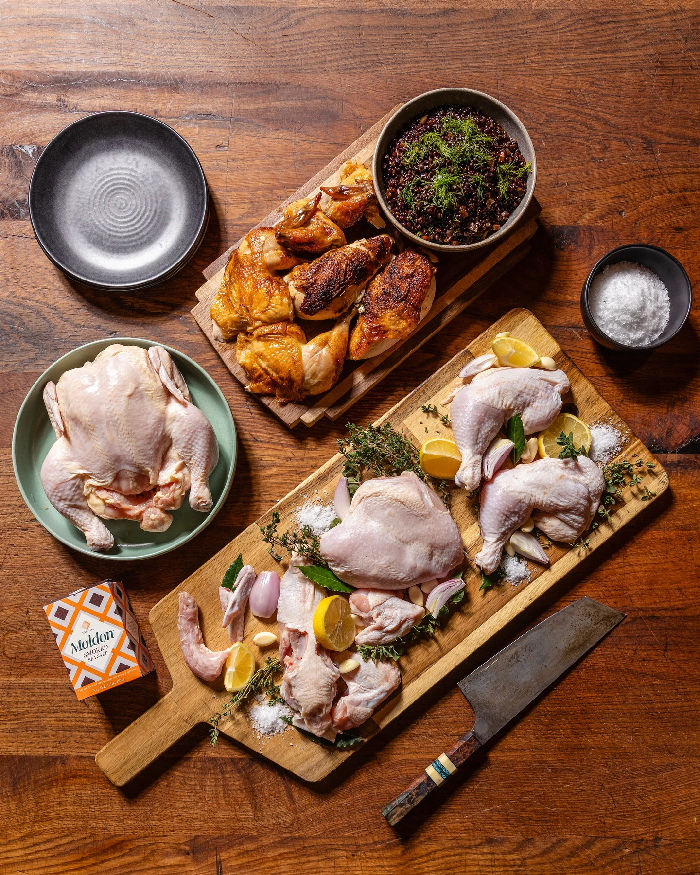 Overhead photo of raw and roasted chicken with lemons, shallots, garlic, thyme, and a chef's knife on a dark wooden table
