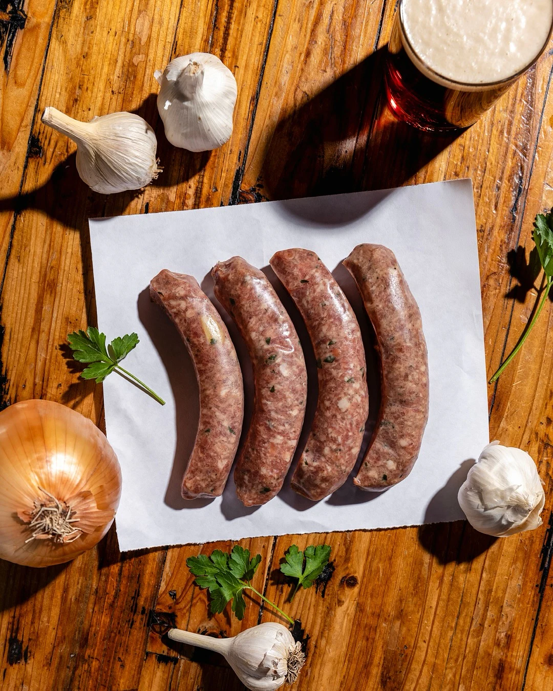Four fresh house-made sausages on butcher paper with garlic, onion, parsley, and a beer
